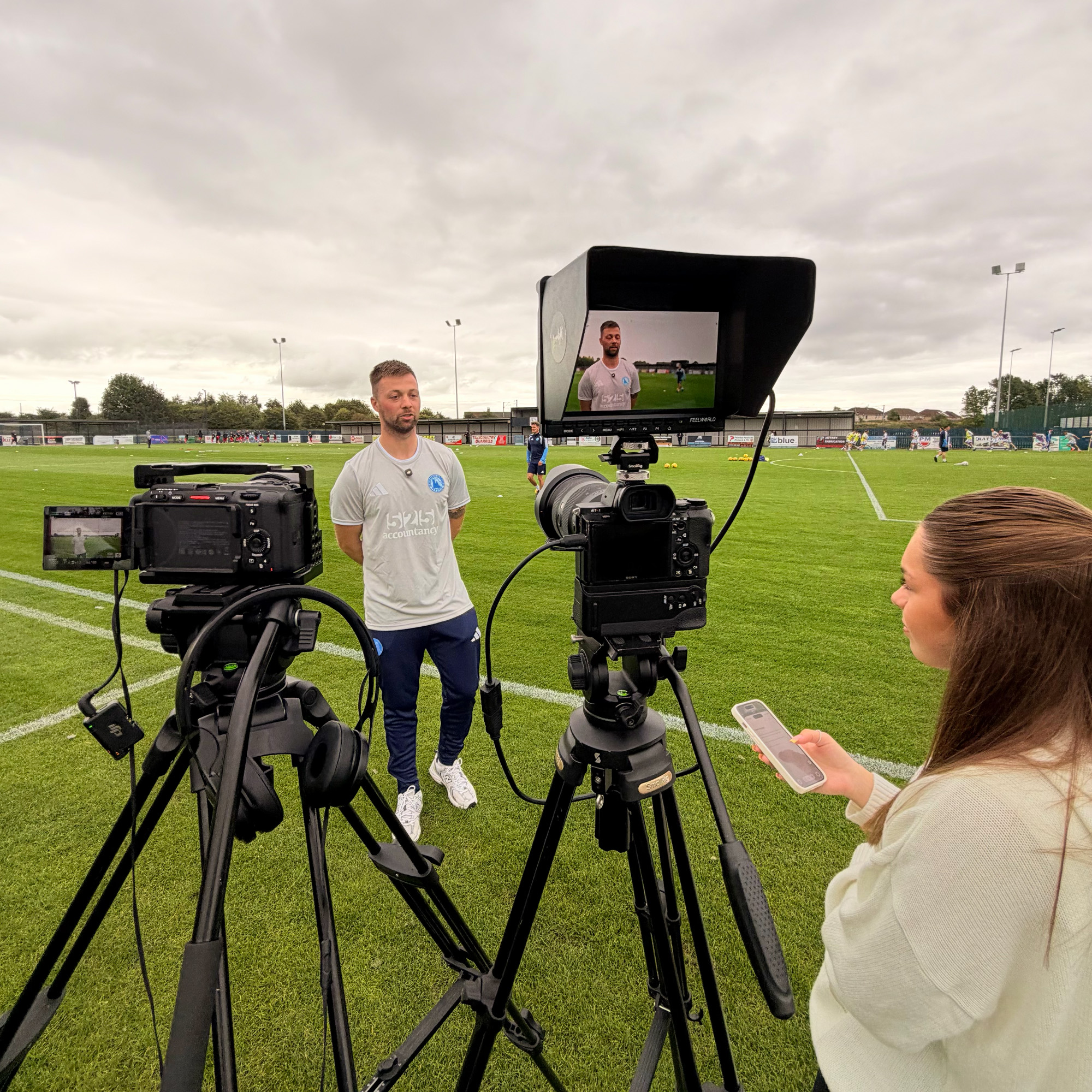 A film crew with two cameras on tripods, a monitor and headphones interviewing a man on a football pitch