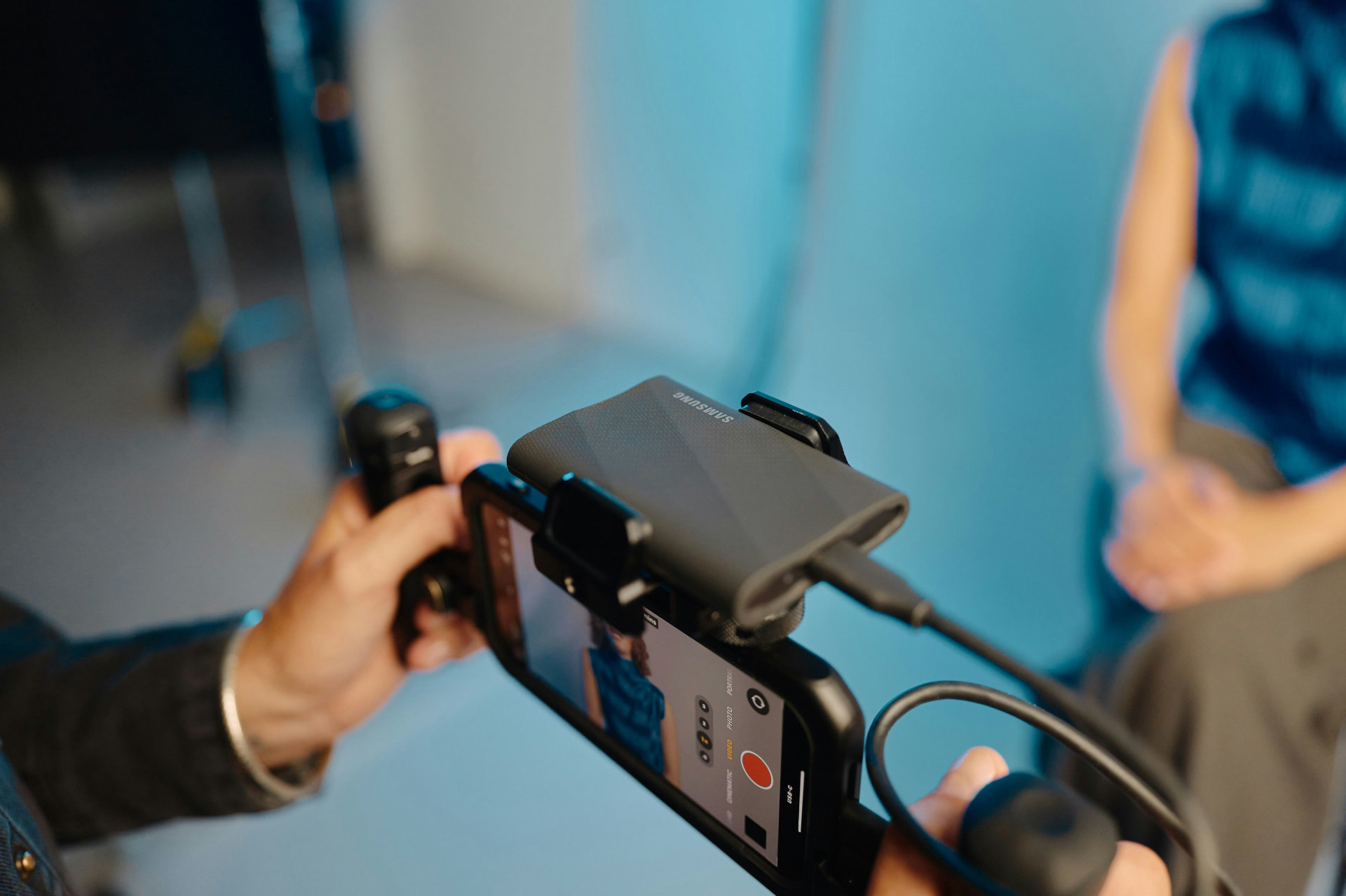 A creator's hands holding a smartphone filming rig with a portable drive mounted on top, in a studio lit with cyan light